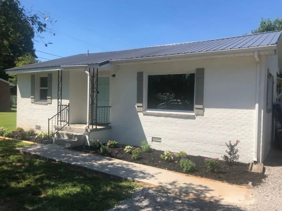 The image showcases the inviting exterior of a newly renovated apartment, featuring a charming front porch framed by lush landscaping and a large window. The space is designed for enjoying quiet mornings with coffee outdoors, relaxing evenings watching the sunset, and gathering with family in a peaceful small-town setting.
