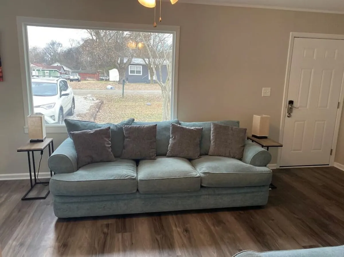 A sunlit living room scene featuring a comfortable sofa, large window with views outside, side tables, and a door. The room feels cozy and is designed for both relaxation and social interactions, with the soft daylight enhancing the inviting atmosphere.