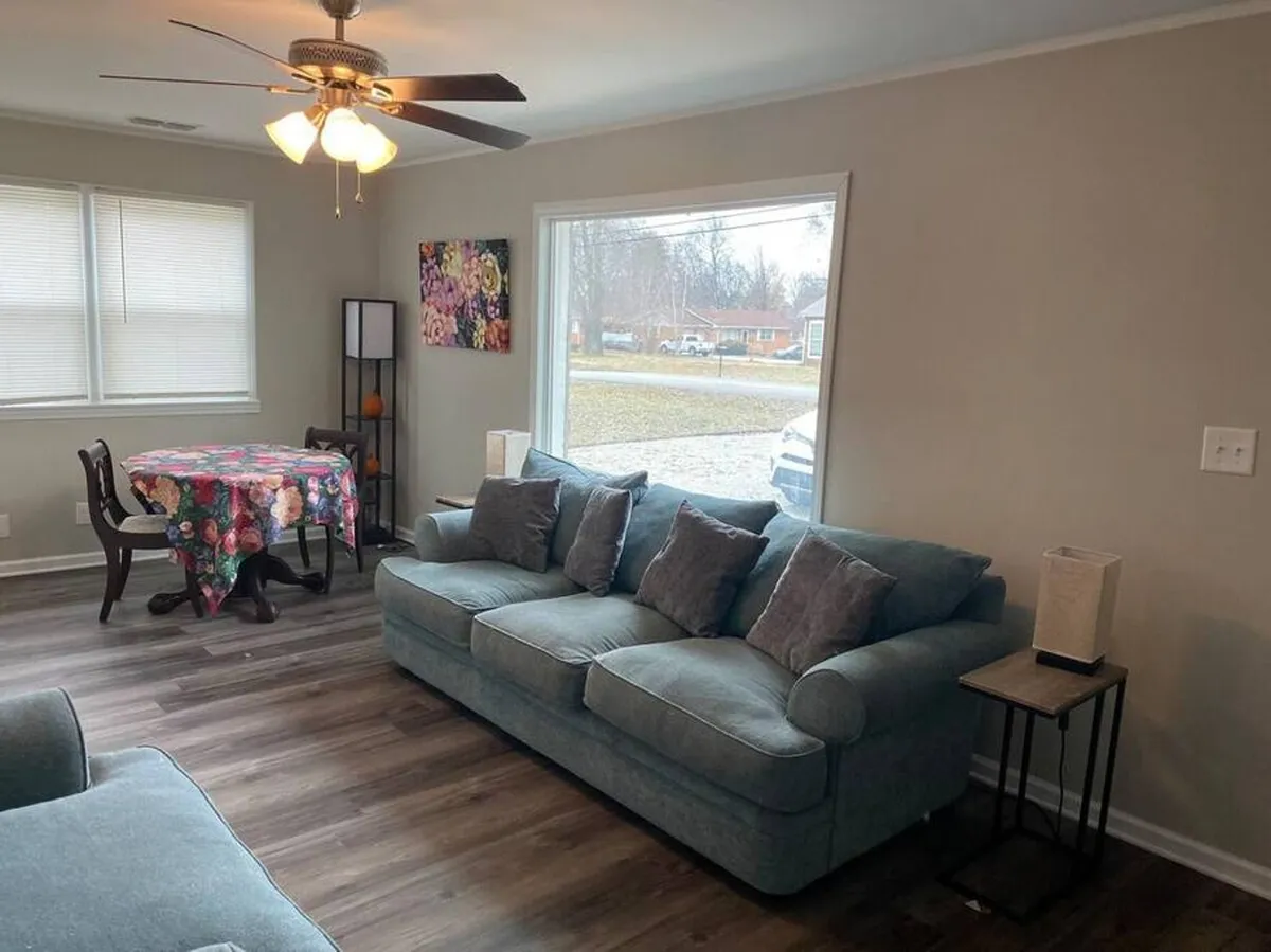 A welcoming living area featuring a plush sofa, a dining table for group meals, and a large window that brings in abundant natural light. The space is arranged for both social gatherings and quiet relaxation, with a view that hints at the outdoors just beyond.