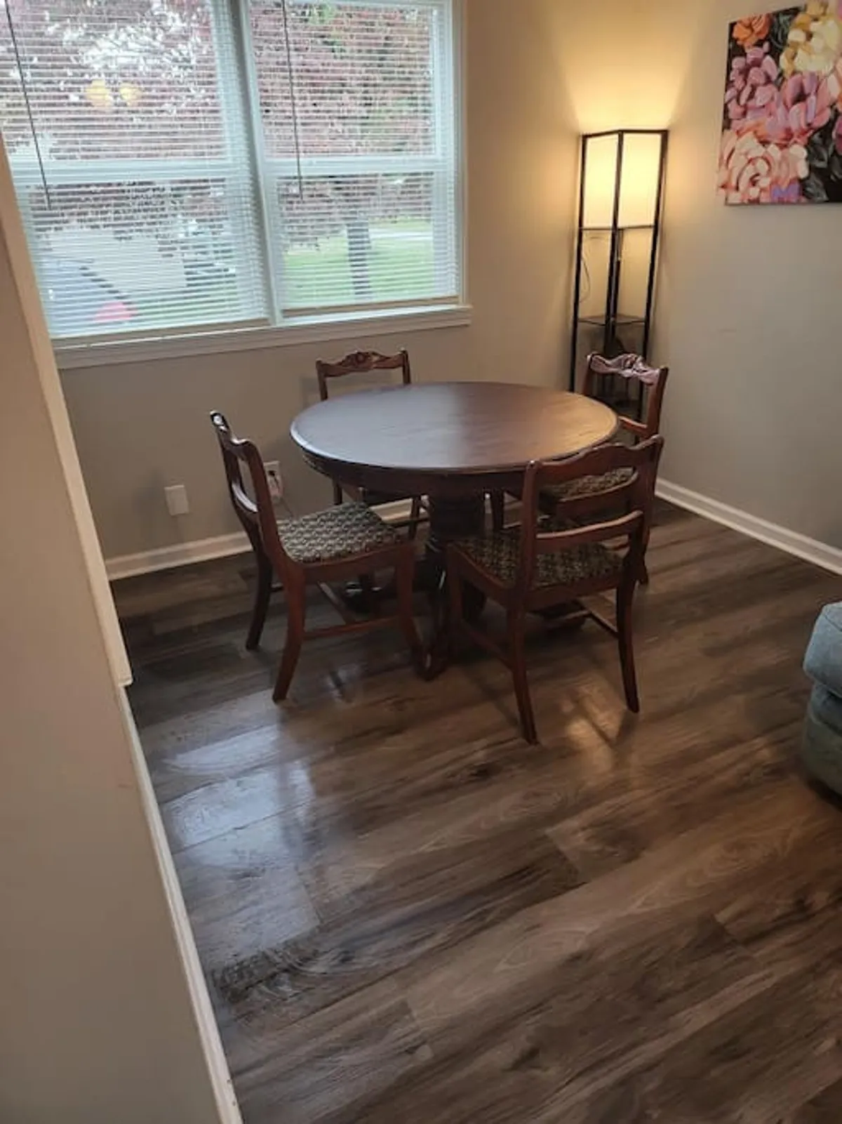 A cozy dining area with a round wooden table surrounded by matching chairs, a window with blinds letting in natural light, and decorative wall art. The setting is warm and inviting, ideal for family meals, conversation, and togetherness.