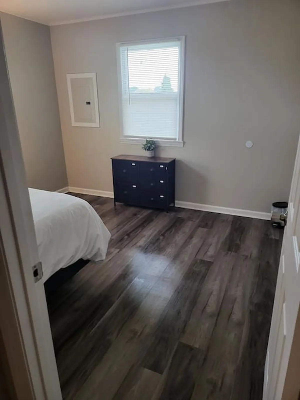 A tranquil bedroom with a neatly made bed facing a window, a dresser nearby, and an inviting reading nook. Soft daylight fills the space, evoking a sense of calm and relaxation.