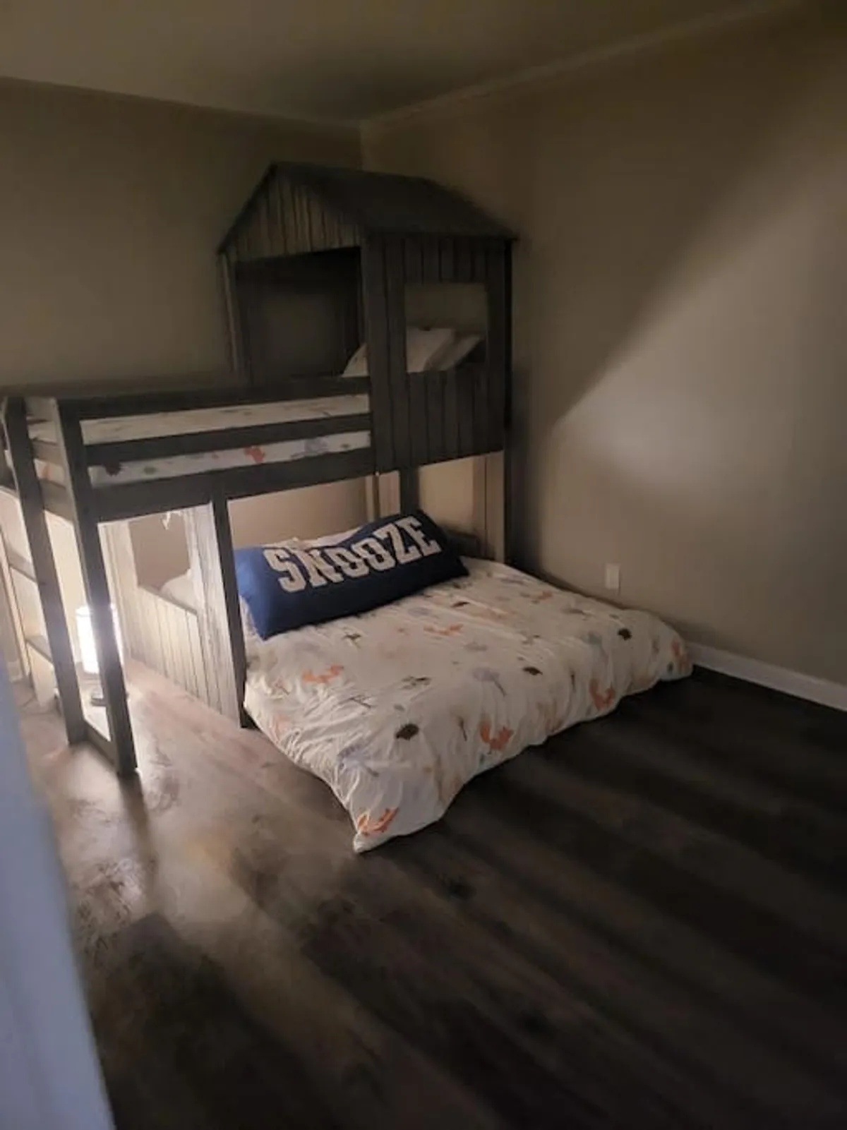 A playful, kid-friendly bunk room featuring a sturdy bunk bed with a whimsical 'SNOOZE' pillow and additional floor bedding. The space is bright and inviting, perfect for imaginative play, sleepovers, or resting between lakeside adventures.